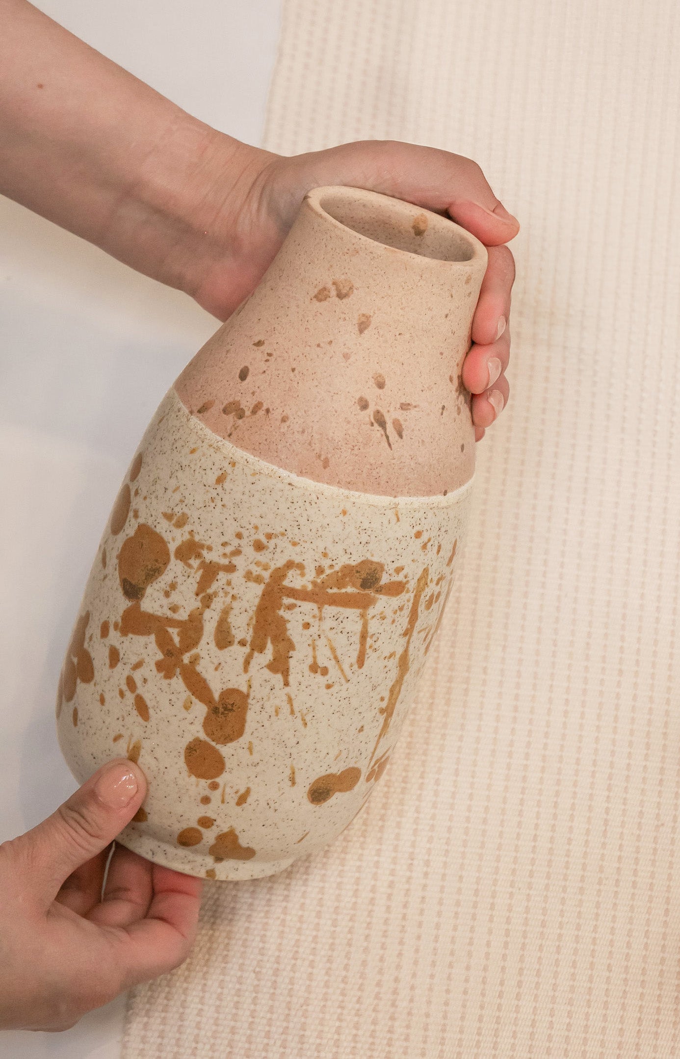 Person holding a ceramic vase with brown speckles on a neutral background